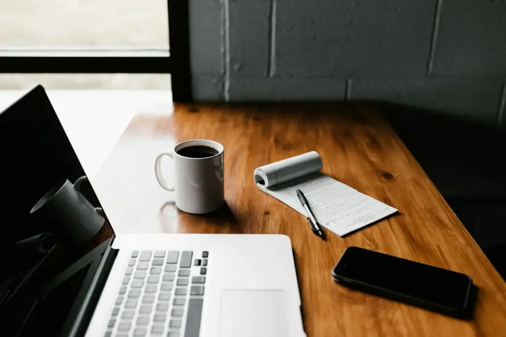 Laptop, coffee mug and notepad on a wooden desk by a window